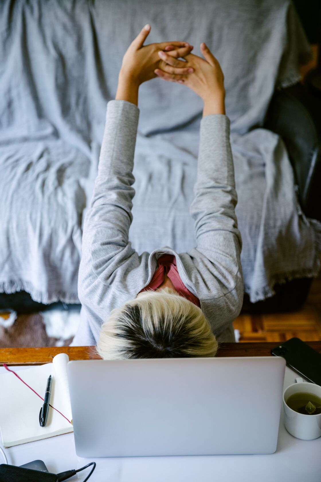 Person at a laptop seen from above, reaching both arms overhead with fingers interlaced