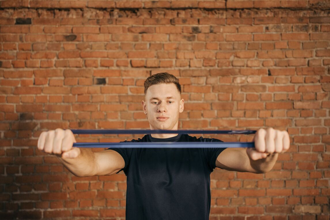 Man holding a resistance band with both hands at chest height in front of a brick wall